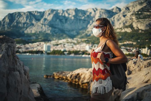 Smiling young female turist with protective mask spending time on seaside during exploring a Mediterranean at corona pandemic. 