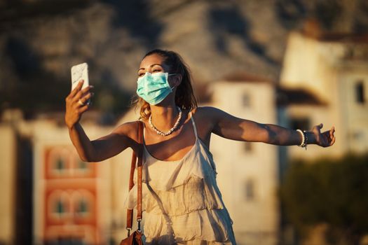 Shot of an attractive young woman wearing a surgical mask and making video call with her smartphone while exploring the wonderful city of Meditteranian.