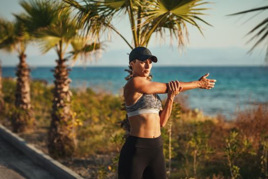 A beautiful young woman is doing stretching exercise at the sea beach in summer sunny day in sunset.