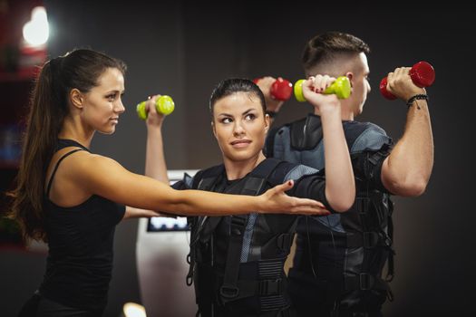 A beautiful young couple are doing exercises with female personal trainer in the gym, dressed in a black EMS suit to stimulate their muscles.