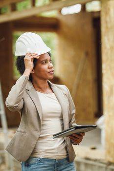 Shot of an African female architect with white helmet using a digital tablet and checking construction site of a new wooden house.