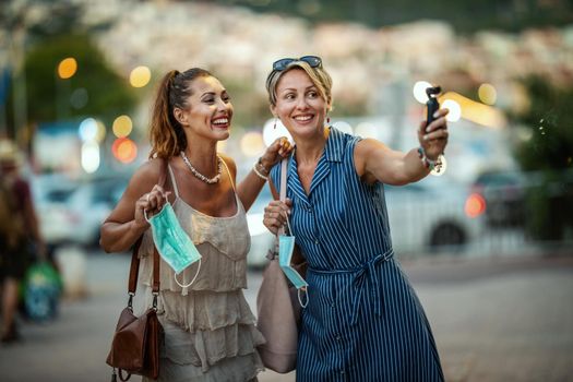 Shot of two attractive female friends who wear a surgical mask and using a digital camera while exploring the wonderful city of Meditteranian.