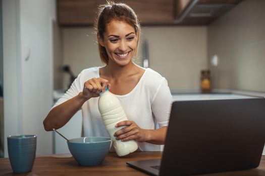 Beautiful young woman is preparing her healthy breakfast in her kitchen and using laptop to make a video chat with someone.