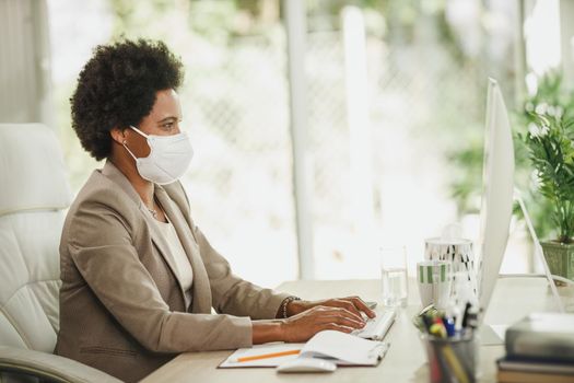 Shot of an African businesswoman with protective N95 mask sitting alone in her office and working on computer during COVID-19 pandemic.