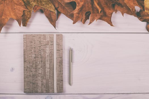 Notebook and pen on wooden table with autumn leaves.Image is intentionally toned.