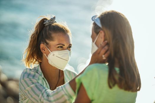 Shot of a happy little girl and her young mom with protective N95 mask spending time on the seashore at corona pandemic. 