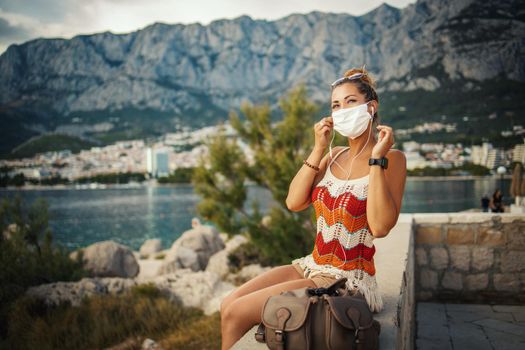 Shot of a happy young woman with protective mask spending time on seaside and listening music during exploring a Mediterranean at corona pandemic. 