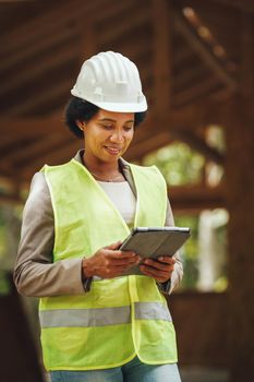 Shot of an African female architect using a digital tablet and checking construction site of a new wooden house. She is wearing protective workwear and white helmet.