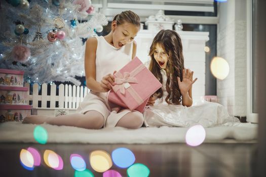 Shot of a two excited little girls opening their Christmas gifts by a white Christmas tree at the home.