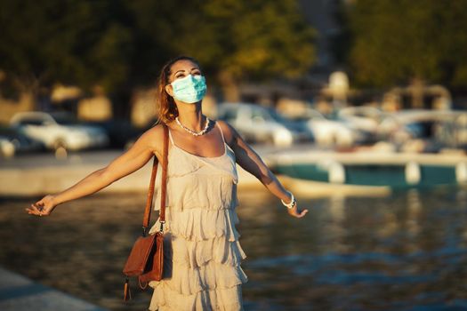 Shot of an attractive young woman wearing a surgical mask while enyoing herself during walk promenade by the sea.