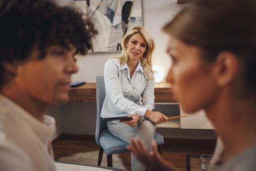 Shot of a couple having a discussion during a counseling session with theirs female psychotherapist while being seated on a sofa inside of a living room.