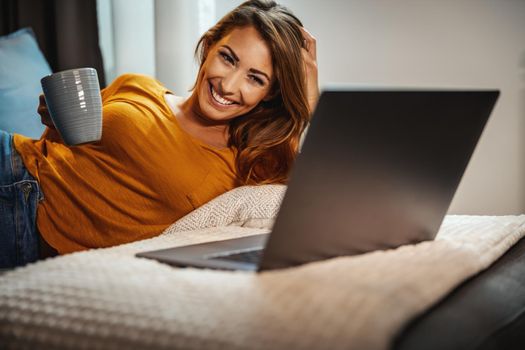 Attractive young woman drinking coffee, lying on the sofa and using her laptop to surfing the net at home.