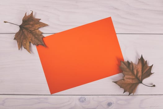 Empty orange paper on wooden table with autumn leaves.Image is intentionally toned