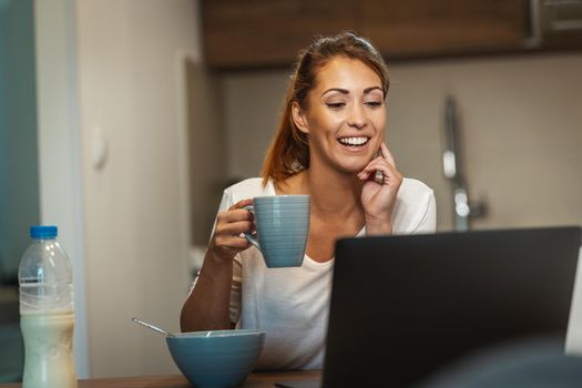 Shot of an cute young woman sitting in the kitchen, enjoying a cup of coffee and using laptop to make a video chat with someone at home.