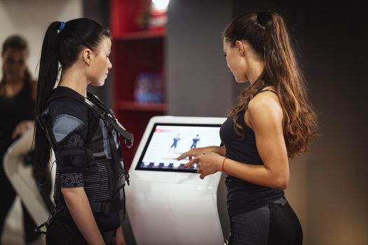 Female personal trainer is teaching a smiling young woman to workout with EMS device in the gym.