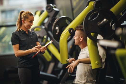 Shot of a muscular young woman in sportswear working out with personal trainer at the gym machine. She is pumping up her chest muscule with heavy weight.