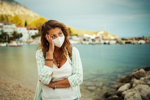 Pensive young woman with protective N95 mask feeling worried while spending time on the seaside during the COVID-19.