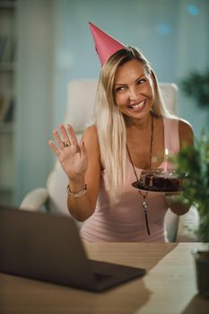 Alone attractive woman have birthday celebration at home during pandemic isolation and have video call with friends. She holding birthday cake with lighted candles.