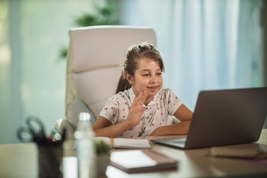 Shot of a cute little girl using her tablet to do a online lesson and waving to teacher over a video call during COVID-19 pandemic.