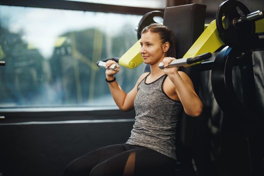 Shot of a muscular young woman in sportswear working out at the gym machine. She is pumping up her shoulder muscule with heavy weight.