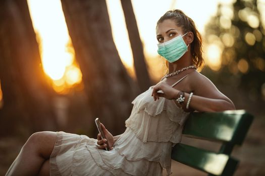 Shot of an attractive young woman wearing a surgical mask and using a smartphone while enyoing herself isolation in the park.