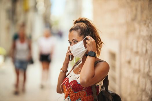 Shot of a happy young woman with protective mask spending time on vacation and exploring a Mediterranean city at corona pandemic. 