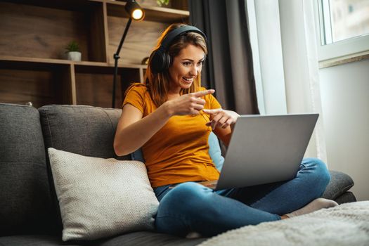 Shot of a attractive young woman sitting cross legged on the sofa and using her laptop and headphones to make a video chat with someone at home.