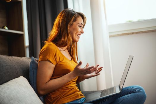 Attractive young woman sitting cross legged on the sofa and using her laptop to make a video chat with someone at home.