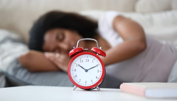 Close-up of red mechanical alarm clock standing on bedside table showing ten minutes to eleven. Young latino woman enjoying sweet dream on background. Wake up concept