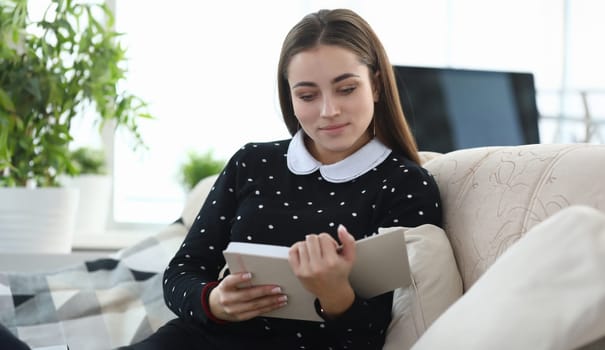 Portrait of young charming lady reading interesting book in living room. Smiling woman looking at notebook with gladness. Relaxation and spare time concept