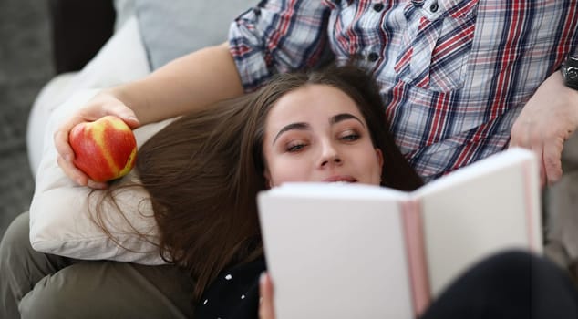 Close-up of young lovely woman laying on partners lap and reading interesting book. Man holding juicy apple. Couple on couch at home. Holiday and relaxation concept