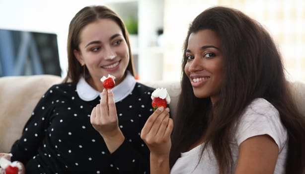 Portrait of two smiling beautiful female holding tasty red berry with whipped cream on top. African-american woman looking at camera with happiness. Friendship and multinational concept