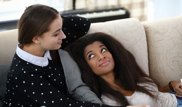 Portrait of best friends laying together on couch. African-american young woman smiling and looking up on another female with tenderness. Friendship concept