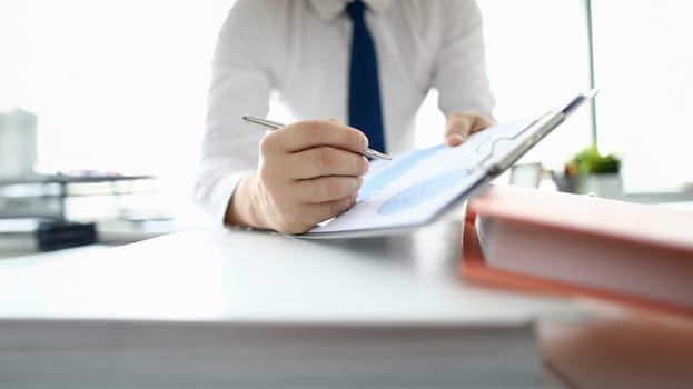 Close-up of businessman hand signing important documents. Manager making annual report and writing down on clipboard. Man in suit sitting in company office. Business and finance concept