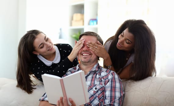 Portrait of two pretty women close both male eyes with hands. Laughing man holding book sitting on couch. Best friends spending fun time together. Friendship and mixed race concept