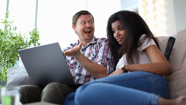 Portrait of surprised man showing something to african-american woman on laptop. Astonished female sitting next to husband on couch. Mixed race family concept