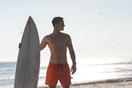 A shirtless man surfer standing on the beach holding a surfboard - looking to the side. Mid shot
