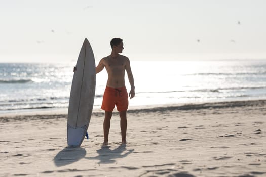 A shirtless man surfer standing on the beach holding a surfboard. Mid shot