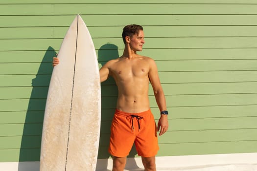 A man standing at the light green wall holding a surfboard. Mid shot