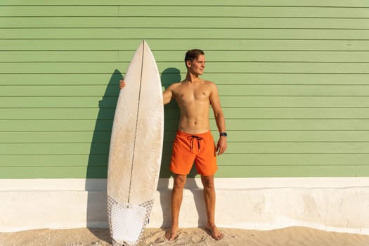 A man with nice body standing at the light green wall holding a surfboard - looking to the side. Mid shot