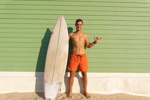 A smiling man with nice body standing at the light green wall holding a surfboard and showing shaka. Mid shot