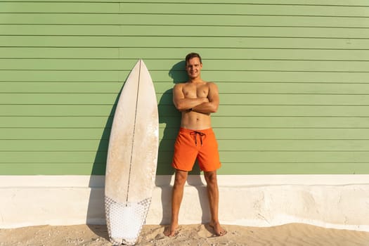A smiling man with nice body standing at the light green with a surfboard. Mid shot