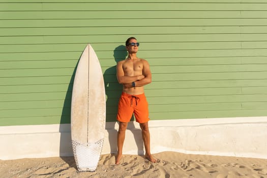 A smiling joyful man with nice body wearing sunglasses standing at the light green with a surfboard. Mid shot