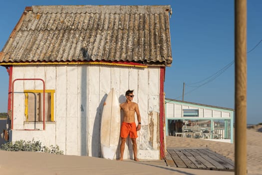 A man with nice athletic body standing at the shore house on the beach with his surfboard - looking to the side. Mid shot