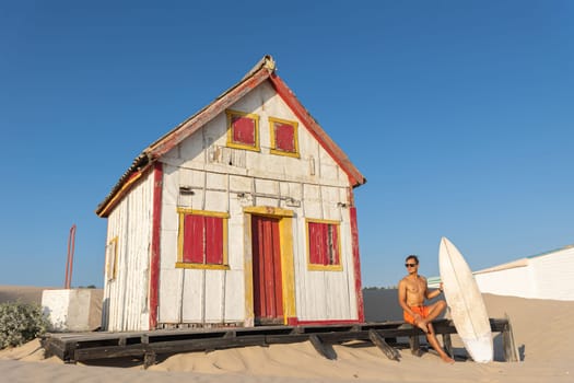 A man with nice athletic body sitting by the shore house holding a surfboard. Mid shot