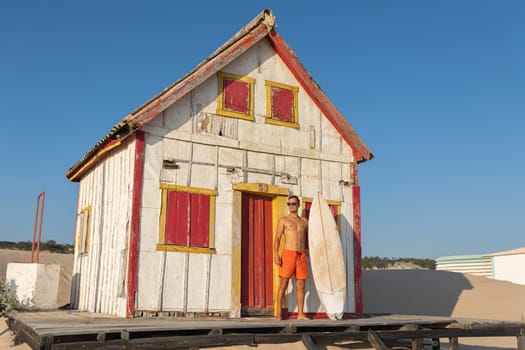 A man with nice athletic body standing at the shore house holding a surfboard. Mid shot