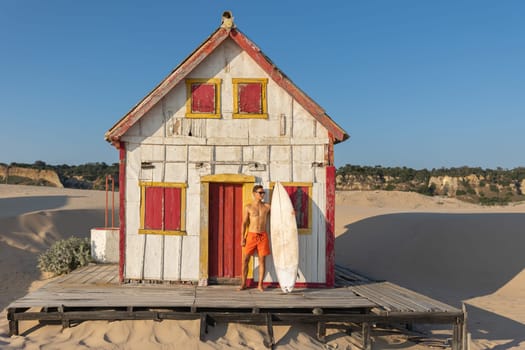 A man with nice athletic body standing at the old seaside house holding a surfboard. Mid shot