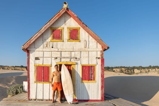 An attractive man with nice athletic body standing at the old seaside house holding a surfboard. Mid shot