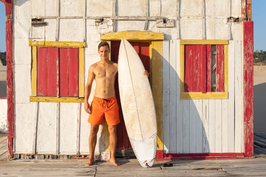 An attractive smiling man with nice athletic body standing at the old seaside house holding a surfboard. Mid shot