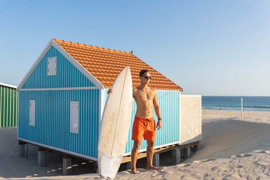 An attractive man with nice athletic body standing at the small seaside building holding a surfboard. Mid shot